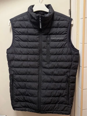 Svart dunväst från Peak Performance - Säljer en supersnygg svart dunväst från Peak Performance med quiltad design och hög krage. Västen har dragkedja framtill och flera praktiska fickor med dragkedja. Knappt använd, jättefint skick. Storlek S, passar även XS. 