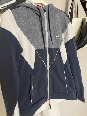 Blå och grå hoodie från BOSS - Snygg hoodie från BOSS i blått, grått och vitt med dragkedja framtill och justerbar huva med snörning. Tröjan har ett sportigt färgblocksmönster och BOSS-logga på bröstet. Perfekt för en avslappnad och trendig stil.