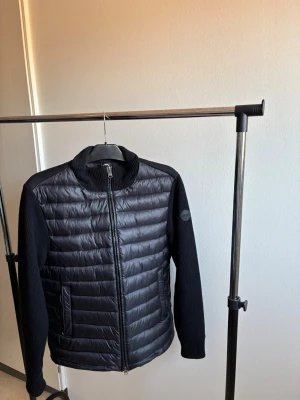 Svart pufferjacka från Colmar - Snygg svart pufferjacka från Colmar med quiltad front i glansigt material och baksidan mjukt samt stickade ärmar och krage. Jackan har dragkedja framtill, två fickor med dragkedja och en diskret logotyp på ärmen. Perfekt för våren 