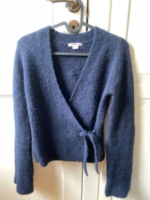 Mörkblå omlottkofta från American Vintage - Mysig mörkblå stickad omlottkofta från American Vintage med långa ärmar och knytband i midjan. Koftan har en djup V-ringning och är tillverkad i ett mjukt, fluffigt material som känns skönt mot huden. Perfekt för lager på lager och en avslappnad look.