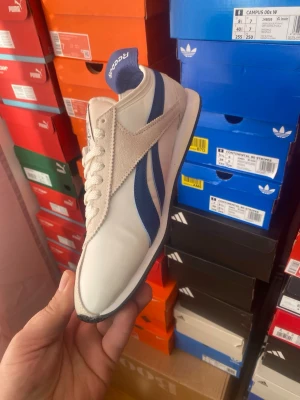 Vita och blå Reebok sneakers - Snygga vita sneakers från Reebok med blå detaljer och klassisk retrodesign. Skorna har låga skaft, vita snören och en ovandel i läder och textil. Perfekta för dig som gillar sportig och stilren look.