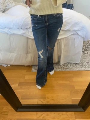 Blå slitna bootcut jeans - Säljer ett par blå bootcut jeans med slitningar och hål på benen. Jeansen har låg midja, klassiska fem fickor och vita kontrastsömmar. Perfekta för en avslappnad och trendig look. Dock vid stygnen har jag sytt eftersom dom sprack , men funkar absolut 