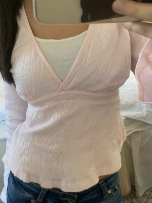 Ljusrosa omlottblus med v-ringning - Söt ljusrosa omlottblus med djup v-ringning och långa ärmar. Blusen har lätt skrynkligt tyg och en figurnära passform som markerar midjan. Perfekt att styla med jeans för en avslappnad men trendig look. man kan även knyta den i ryggen. 