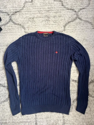 Marinblå kabelstickad tröja Morris - Snygg marinblå kabelstickad tröja från Morris i merceriserad bomull. Tröjan har rund halsringning med röd detalj på insidan och en liten röd logotyp broderad på bröstet. Perfekt för dig som gillar klassisk stil med en twist.