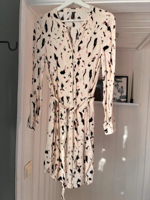 Mönstrad kortklänning med knappar - Säljer en ljus kortklänning med svart och rosa abstrakt mönster. Klänningen har lång ärm, knappar framtill, v-ringning och ett matchande bälte i midjan. Perfekt för dig som gillar en feminin och avslappnad stil.