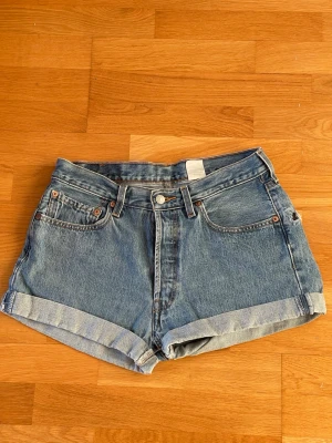 Levi's 501 blå denimshorts hög midja W33 - Klassiska blå denimshorts från Levi's 501 med hög midja och uppvikta benslut. Storlek W33. Femficksmodell med knappgylf och tydliga sömmar. Levi's ikoniska läderpatch bak och röd etikett på ena bakfickan. Perfekta till sommaren!
