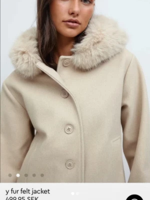 Beige kappa med fuskpäls från Gina Tricot - Stilren beige kappa från Gina Tricot med stor fluffig fuskpäls runt kragen. Kappan har knäppning framtill med stora knappar, raka ärmar och två fickor. Materialet är filtad ullmix som ger en mjuk och lyxig känsla. Perfekt för kalla dagar.