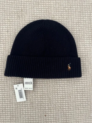 Marinblå ribbstickad mössa Polo Ralph Lauren - Säljer en marinblå ribbstickad mössa från Polo Ralph Lauren med klassisk uppvikt kant och den ikoniska broderade loggan framtill. Mössan är tillverkad i mjukt material och passar perfekt för kyliga dagar.