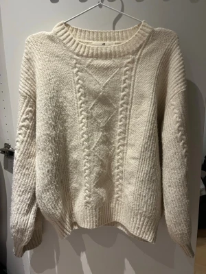 Vit kabelstickad tröja från H&M - Mysig vit kabelstickad tröja från H&M med grov struktur och ribbade muddar. Tröjan har ett klassiskt flätmönster framtill, rund halsringning och långa ärmar. Perfekt för kalla dagar och enkel att matcha med jeans eller kjol.