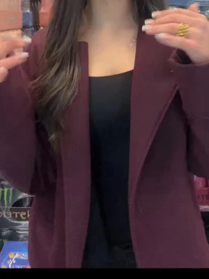 Vinröd kavaj med enkel design - Stilren vinröd kappa med rak passform och breda slag. Kappan är utan synliga knappar och har en mjuk struktur, perfekt att matcha med en enkel topp. Materialet ser ut att vara en blandning av polyester och bomull.