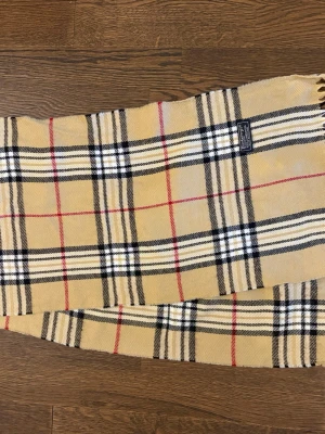 Beige Burberry Halsduk - Klassisk beige halsduk från Burberry i ull med det ikoniska rutmönstret i svart, vitt och rött. Hör av dig vid funderingar!
