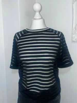 Randig svartvit mesh t-shirt - Snygg svart och vit randig t-shirt i meshmaterial. T-shirten har korta ärmar och rund halsringning. Har ett pytte litet hål vid bakre delen av kragen som knappt syns