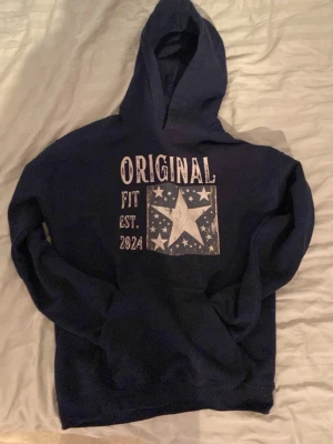 Mörkblå hoodie med stjärntryck - Mörkblå hoodie från Original Fit med stor vit stjärna och texten 'ORIGINAL FIT EST. 2021' på bröstet. Hoodien är typ oanvänd. Hoodien har huva och magficka framtill. Perfekt för en avslappnad och cool stil. Materialet känns mjukt och bekvämt.  (HOODIEN HAR INGA SNÖREN OCH DÄRFÖR HAR JAG SÄNKT PRISET)