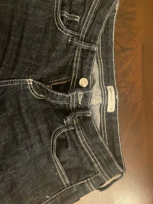 Mörkblå jeans från Ginatricot - Snygga mörkblå jeans från Gina med kontrasterande vita sömmar och klassisk femficksdesign. Jeansen har en knappgylf och är tillverkade i ett mjukt denimtyg som ger en bekväm passform. Perfekta för en avslappnad och trendig look.