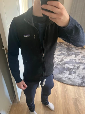 Svart dunväst från Patagonia - Snygg svart dunväst från Patagonia med dragkedja framtill och bröstficka med logga. Västen har en klassisk passform och är perfekt att slänga över en hoodie eller tröja för extra värme. Enkel och stilren design som funkar till det mesta.