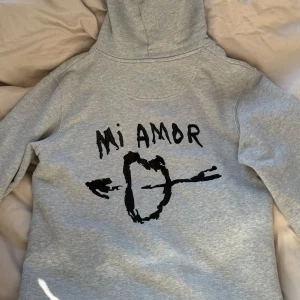 Grå hoodie med tryck 'Mi Amor' - Säljer en grå hoodie med huva och känguruficka. Orginalpris 1400kr. På ryggen finns ett svart tryck med texten 'Mi Amor' och ett hjärta genomborrat av pilar. Hoodien har en avslappnad passform och är tillverkad i mjukt sweatshirtmaterial.