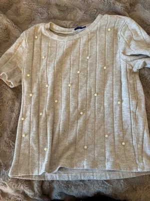 Beige ribbad topp med pärlor - Trendig beige topp med ribbad struktur och dekorativa pärlor framtill. Kortärmad modell med rund halsringning, perfekt för en stilren och söt look. Materialet känns mjukt och bekvämt mot huden.