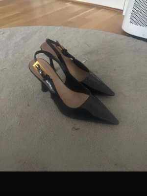 Svarta lackpumps med öppen häl - Säljer ett par svarta pumps i lack med spetsig tå och öppen häl. Skorna har smal klack och justerbar rem runt hälen. Snygg och klassisk modell som ger en elegant look. Perfekta till både jeans och klänning.