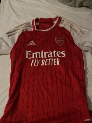 Arsenal matchtröja röd/vit Adidas - Knappt använd. Snygg Arsenal matchtröja från Adidas i rött med vita ärmar och gulddetaljer. Tröjan har klubbmärke och Adidas-logga på bröstet samt 'Emirates Fly Better' tryckt framtill. Tillverkad i lätt och ventilerande material, perfekt för fotboll eller träning.