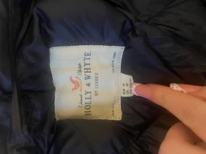 Mörkblå pufferjacka Holly & Whyte - Snygg mörkblå pufferjacka från Holly & Whyte by Lindex med quiltat mönster och guldfärgade knappar. Jackan har hög krage och en klassisk, puffig siluett som håller dig varm under kalla dagar. Perfekt för dig som vill ha både stil och funktion.