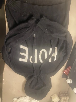 Svart hoodie med HOPE-tryck - Säljer en svart hoodie med vit text 'HOPE' på bröstet. Hoodien har dragsko i huvan och ribbade muddar vid ärmar och nederkant. Materialet är stickat och känns mjukt och bekvämt. Perfekt för dig som gillar en enkel men snygg stil.