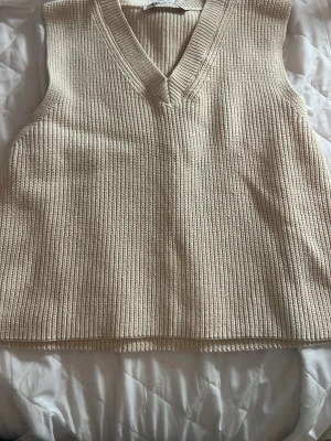 Beige ribbad stickad väst från Zara - Säljer en beige stickad väst från Zara med ribbad struktur och v-ringad hals. Perfekt att styla över en skjorta eller t-shirt för en trendig lager-på-lager-look. Västen har en loose passform och är superenkel att matcha med olika outfits.