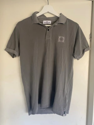 Grå pikétröja från Stone Island - Snygg grå pikétröja från Stone Island med klassisk krage, korta ärmar och knappslå framtill. Tröjan har den ikoniska Stone Island-loggan broderad på bröstet och är tillverkad i ett mjukt bomullsmaterial. Perfekt till jeans eller shorts.