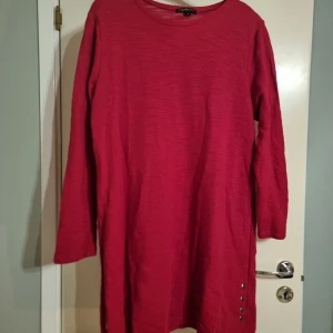 Röd långärmad klänning med knappar - En enkel röd klänning med lång ärm och rund halsringning. Klänningen har dekorativa knappar på sidan nedtill som ger en snygg detalj. Passar perfekt för dig som gillar stilrena plagg med en twist.
