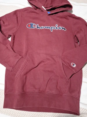 Vinröd hoodie från Champion - Säljer en vinröd hoodie från Champion med broderad logga på bröstet och liten logga på ärmen. Klassisk känguruficka framtill och mjukt material. Perfekt för en avslappnad och sportig stil.
