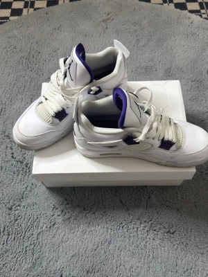Vita Air Jordan 4 med lila detaljer - Säljer ett par vita Air Jordan 4 sneakers med lila detaljer och klassisk Jumpman-logga på plösen. Skorna har meshpaneler, vita skosnören och en robust sula. Insidan är fodrad i lila och materialet är en mix av läder och textil. Perfekta för dig som vill sticka ut med stil.