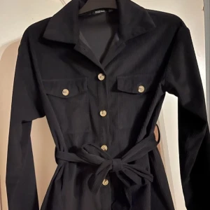 Svart manchesterskjorta med knappar - Stilren svart skjorta från Boohoo i mjuk manchester med långa ärmar, markerad midja med knytband och snygga knappar framtill. Två bröstfickor med knappdetaljer och klassisk krage ger en cool touch.