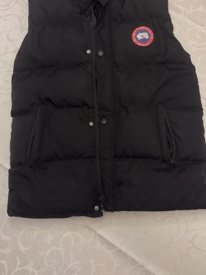 Svart dunväst från Canada Goose - Snygg svart dunväst från Canada Goose med klassisk logotyp på bröstet. Västen har dragkedja och tryckknappar framtill, två fickor med dragkedja och en innerficka. Perfekt för lager på lager och håller dig varm under kyliga dagar.