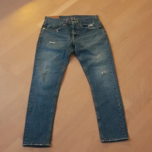 Blå dondup jeans - Ett par blå jeans med rak passform och slitna detaljer på både fram- och baksida. Jeansen har klassisk femficksmodell, knappgylf och råa kanter vid bensluten. Perfekta för en avslappnad och trendig look.