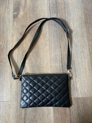 Svart quiltad axelväska med kedjedetaljer - Snygg svart axelväska i quiltat mönster med justerbar axelrem och guldfärgade detaljer. Väskan har dragkedja upptill och slät baksida. Perfekt för att bära det viktigaste och ger en trendig touch till din outfit.