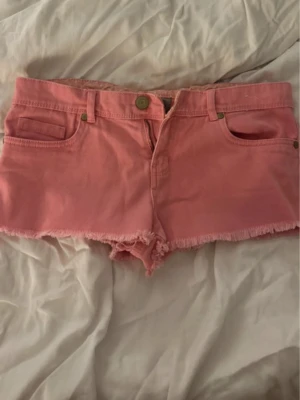 Rosa jeansshorts med fransig kant - Säljer ett par ljusrosa jeansshorts med fransig kant nertill. Shortsen har fem fickor, bälteshällor och knappstängning framtill. Perfekta för sommaren och ger en avslappnad vibe.