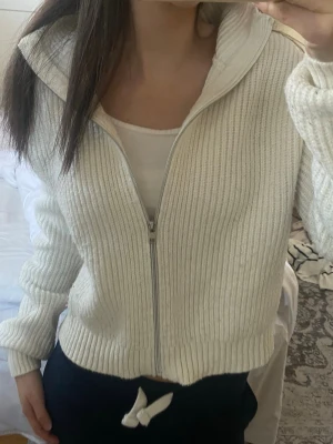 Vit ribbad ziptröja med krage - Säljer en vit ribbstickad ziptröja med hög krage och dragkedja framtill. Tröjan har långa ärmar och en croppad passform som ger en avslappnad look. Perfekt att slänga på sig över ett linne eller t-shirt.