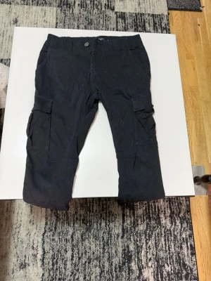 Svarta cargobyxor med fickor - Säljer ett par svarta cargobyxor med klassiska stora sidofickor och knappstängning. Byxorna har en rak passform och är tillverkade i ett slitstarkt material som passar perfekt för dig som gillar en avslappnad och cool stil.