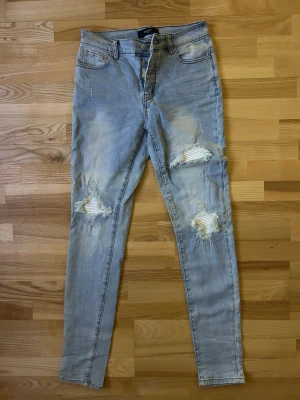Ljusa slitna skinny jeans från Amiri  - Säljer ett par ljusblå skinny jeans från Amiri med slitningar och hål på båda benen. Jeansen har klassisk femficksmodell, hög midja och dragkedja med knapp. Perfekta för en avslappnad och trendig look.