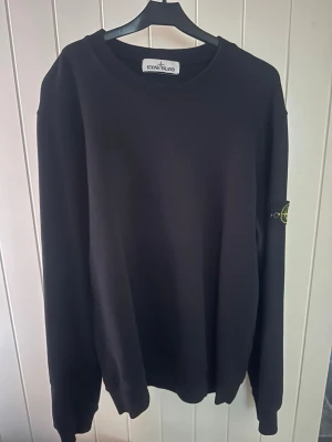 Svart sweatshirt från Stone Island - Svart sweatshirt från Stone Island med klassisk rund halsringning och ikonisk logopatch på vänster ärm. Tröjan har en enkel, stilren design och är tillverkad i mjukt material som passar perfekt till streetwear-looken.