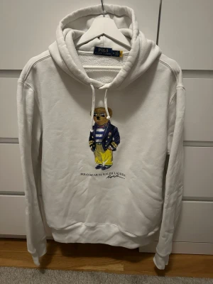 Vit hoodie Polo Bear Ralph Lauren - Vit hoodie från Polo Ralph Lauren med den ikoniska Polo Bear-figuren tryckt på bröstet. Hoodien har dragsko i huvan och ribbade muddar. Tillverkad i mjukt material som känns skönt mot huden. Perfekt för dig som gillar klassisk och stilren streetwear.