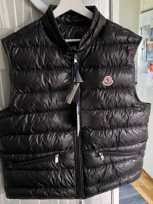 Moncler gui väst storlek 5 - Snygg svart dunväst från Moncler med glansig finish och klassisk logga på bröstet. Västen har två dragkedjeförsedda fickor framtill och hög krage. Tillverkad i nylon med fyllning av dun och fjäder. Perfekt för lager-på-lager och streetstyle.