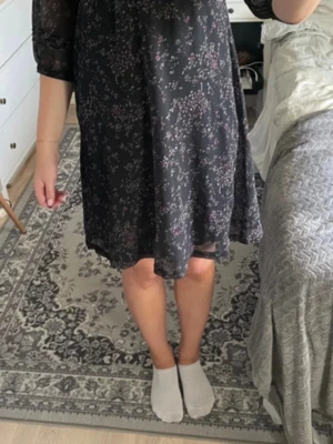 Svart klänning med lila blommor  - Säljer en svart kortklänning med småblommigt mönster i lila och rosa toner. Klänningen har trekvartsärmar i transparent material och en lös, flowy passform. Perfekt för dig som gillar en enkel men söt stil.