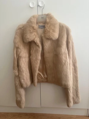 Beige pälsjacka  från Tessie - Snygg beige pälsjacka från Tessie med klassisk krage och rak passform. Jackan har mjukt foder och stängs framtill. Perfekt för dig som vill ha en varm och trendig look under vintern