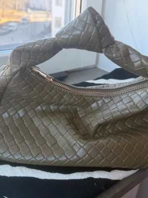 Olivgrön flätad handväska - Trendig olivgrön handväska med flätad design och vridet handtag. Väskan har en snygg silvrig dragkedja och är gjord i mjukt konstläder. Perfekt accessoar för dig som gillar stilrena detaljer och modern look.