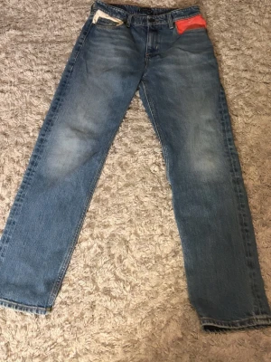 Blå raka jeans med färgdetaljer - Säljer ett par klassiska blå jeans med raka ben och coola färgdetaljer vid fickorna fram, en vit och en röd. Jeansen har fem fickor och normal midja. Perfekta för en avslappnad och trendig look.