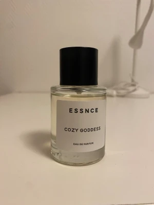 ESSNCE Cozy Goddess Eau de Parfum - Minimalistisk parfymflaska från ESSNCE med doften Cozy Goddess. Flaskan är genomskinlig med svart cylinderformad kork och vit etikett med svart text. Modern och stilren design som passar perfekt på sminkbordet.