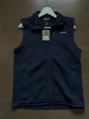 Helt ny Mörkblå fleeceväst från Patagonia - Mörkblå väst från Patagonia i mjuk fleece med hel dragkedja framtill och två sidofickor med dragkedja. Klassisk ståkrage och diskret logga på bröstet. Perfekt lager för kyliga dagar och enkel att matcha med olika outfits.