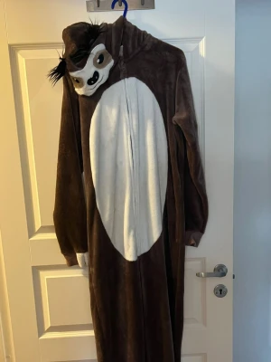Brun sengångare onesie med huva - Mysig brun onesie i plysch med vit mage och huva som har ett sengångaransikte och svarta detaljer. Dragkedja framtill och långa ärmar. Perfekt för maskerad eller cosplay när du vill vara extra chill.