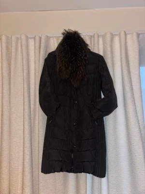 Svart lång dunjacka Rockandblue - Snygg svart medellång dunjacka från Rockandblue med avtagbar pälskrage i äkta päls. Jackan har dragkedja och knappar framtill samt två fickor med dragkedja. Perfekt för kalla vinterdagar och har en stilren, figurnära passform.