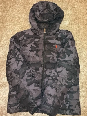 Svart camo pufferjacka från Polo Ralph Lauren - Säljer en svart pufferjacka med grått camomönster från Polo Ralph Lauren. Jackan har huva, dragkedja framtill och den klassiska röda loggan på bröstet. Perfekt för kalla vinterdagar och riktigt snygg streetstil.